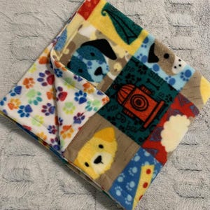 May include: A colorful dog-themed blanket with a patchwork design. The blanket features various dog faces, paw prints, and a fire hydrant on a soft, fleece material. The reverse side displays a pattern of colorful paw prints.