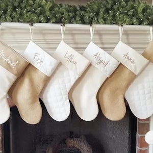 May include: Six personalised Christmas stockings with burlap and white fabric, hanging on a white mantelpiece. The stockings are embroidered with the names Michael, Erin, Lillian, Abigail, Anna, and Bella. A white bone-shaped stocking with the name Buster is hanging below the others.