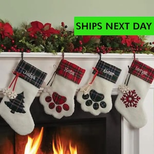 May include: Four personalized Christmas stockings hanging above a fireplace. Each stocking is white with a plaid cuff and features a name, a Christmas tree, paw prints, or a snowflake design. A garland of red berries and flowers decorates the mantel.