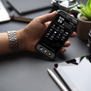 May include: A person's hand holding a black smartphone with a dark mode interface. The screen displays a calendar with the date "SUN 25" and a grid of app icons, including social media apps like TikTok, Twitter, and Instagram. The phone is on a black surface with a tablet and a stylus.