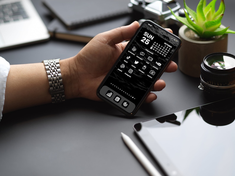 May include: A person's hand holding a black smartphone with a dark mode interface. The screen displays a calendar with the date "SUN 25" and a grid of app icons, including social media apps like TikTok, Twitter, and Instagram. The phone is on a black surface with a tablet and a stylus.