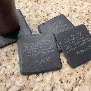 May include: Four black slate coasters with inspirational quotes engraved in white. The quotes are from Romans 8:28, Philippians 4:6, and two other verses.