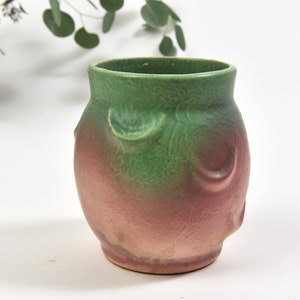 May include: A ceramic vase with a rounded body and a wide opening. The vase features a gradient of colours, transitioning from green at the top to a reddish-pink at the base. Decorative, curved patterns adorn the sides. A decorative piece for the home.