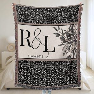 May include: A black and white woven throw blanket with a floral pattern and fringe. The centre features the letters "R & L" and the date "7 June 2019" with a floral design. The blanket is held up by a person.