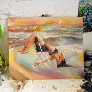 May include: An oil painting depicts a person reclining in a chair on a beach, using a phone. The artwork features a sunset with warm colors, a crescent moon, and the ocean. The person is wearing a black top and shorts.
