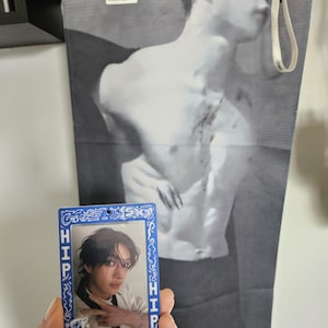 Puede incluir: Una tarjeta fotográfica azul y blanca con la palabra "HIP" en los lados, que presenta a una persona con cabello oscuro. Un póster en blanco y negro de una persona sin camisa está en el fondo.
