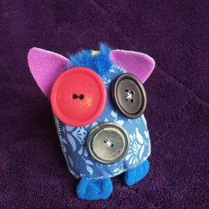 May include: A blue fabric toy with pink ears and blue feet. The toy has three buttons for eyes: one red, one black, and one brown.