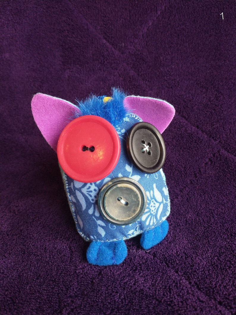 May include: A blue fabric toy with pink ears and blue feet. The toy has three buttons for eyes: one red, one black, and one brown.
