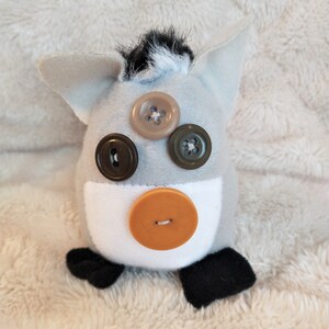 May include: A grey and white plush toy with black ears and a black tail. The toy has brown, orange and green button eyes.