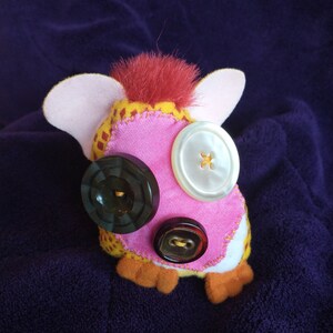 May include: A pink and yellow plush toy with red hair and three buttons for eyes. The buttons are black, white, and brown.