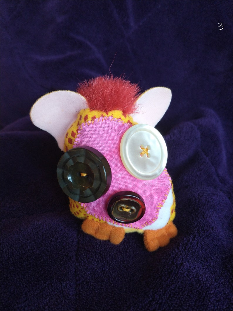 May include: A pink and yellow plush toy with red hair and three buttons for eyes. The buttons are black, white, and brown.