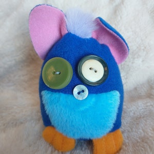 May include: Blue and turquoise plush toy with large green, white and black button eyes. The toy has pink ears and orange feet.