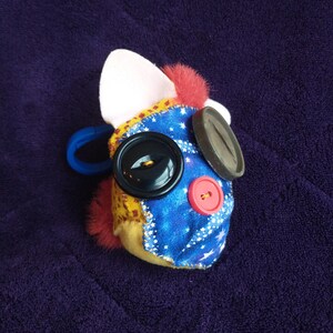 May include: A plush toy with a blue and yellow fabric face, featuring a large black button eye, a red button nose, and a black button on the side. The toy has a white ear and a pink fur patch.