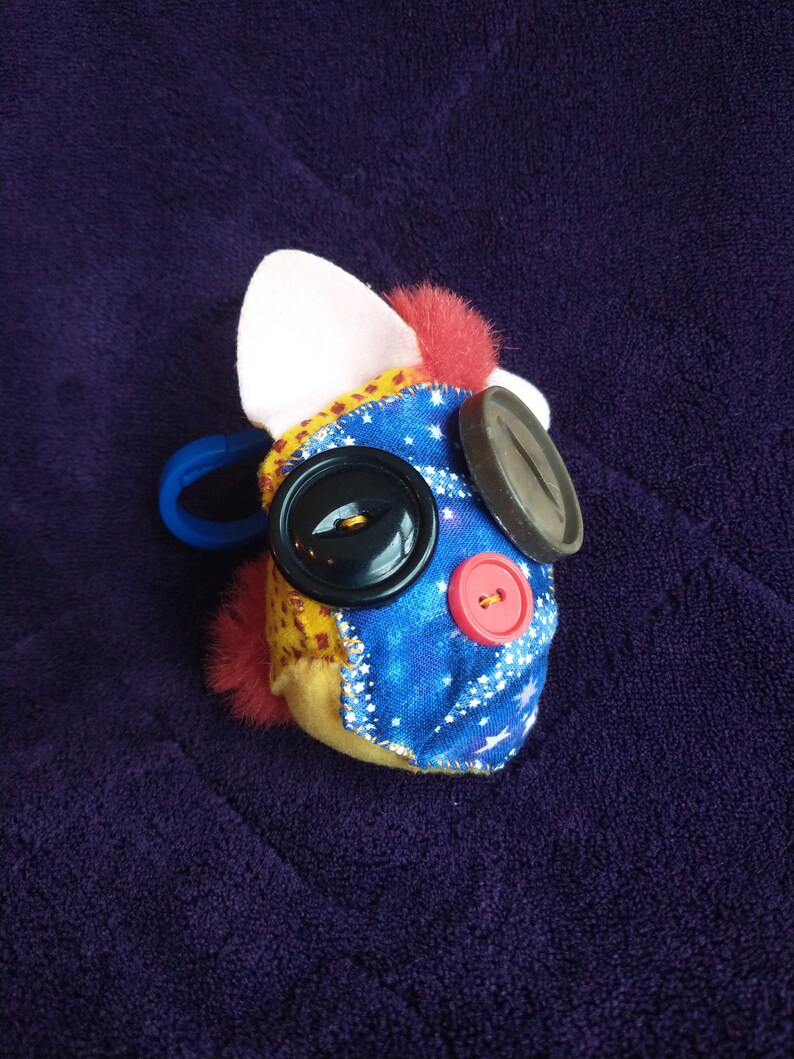May include: A plush toy with a blue and yellow fabric face, featuring a large black button eye, a red button nose, and a black button on the side. The toy has a white ear and a pink fur patch.