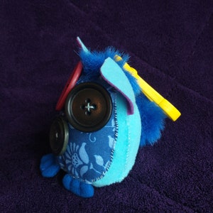 May include: A blue and purple plush toy with a yellow felt wing, blue felt ears, and three black buttons for eyes. The toy is sitting on a purple fabric surface.