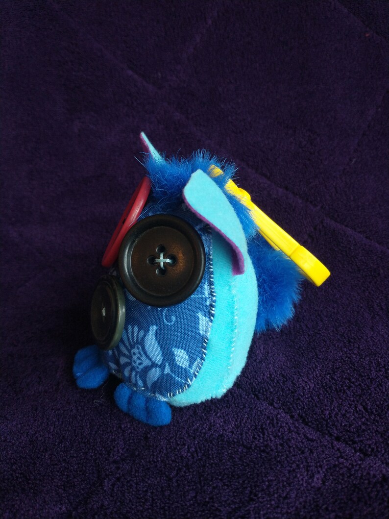 May include: A blue and purple plush toy with a yellow felt wing, blue felt ears, and three black buttons for eyes. The toy is sitting on a purple fabric surface.