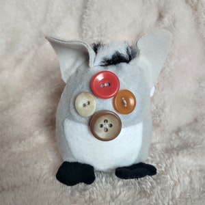 May include: A grey plush toy with black fur on top and four buttons on the front. The buttons are red, brown, beige, and orange.