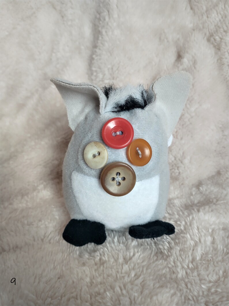 May include: A grey plush toy with black fur on top and four buttons on the front. The buttons are red, brown, beige, and orange.