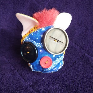 May include: A handmade plush toy with a blue fabric covered body, a red fuzzy head, and white ears. The toy has three buttons for eyes and a mouth. The buttons are black, grey, and red.