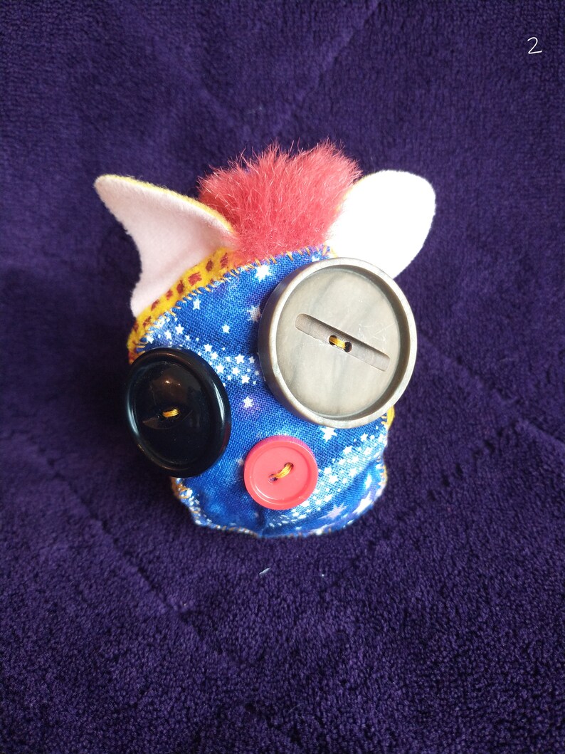 May include: A handmade plush toy with a blue fabric covered body, a red fuzzy head, and white ears. The toy has three buttons for eyes and a mouth. The buttons are black, grey, and red.
