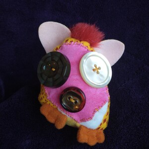 May include: A pink and yellow plush toy with red fur on top. The toy has three buttons for eyes and a mouth. The buttons are brown, white, and black.
