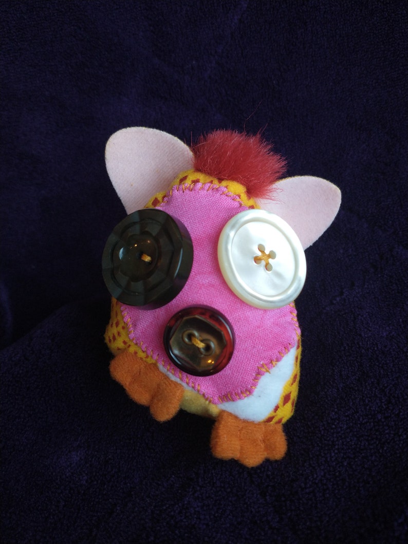 May include: A pink and yellow plush toy with red fur on top. The toy has three buttons for eyes and a mouth. The buttons are brown, white, and black.