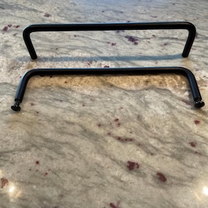 May include: Two black metal cabinet pulls with a rectangular shape. Each pull has a curved design with a screw on each end for installation. The pulls are shown on a speckled, light-colored surface.