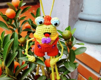 May include: A crocheted toy in the shape of a bug with yellow, orange, and purple accents. The toy is sitting on a potted plant with green leaves and small orange fruits.