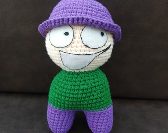May include: A crocheted plush toy with a purple hat, green shirt, and purple legs. The toy has a white face with large eyes, a wide smile, and a black outline.