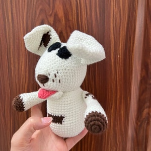 May include: White crocheted dog plush toy with brown spots and a pink tongue sticking out. The toy has black eyes and a brown nose.
