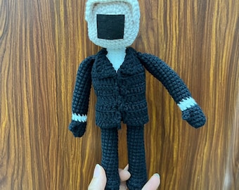 May include: A crocheted doll wearing a black suit with white accents. The doll has a white crocheted head with a black square for a face.
