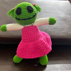 May include: A crocheted doll with a green head, white arms, and a pink dress. The doll has black eyes and a smiling mouth.