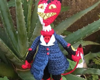 May include: A crocheted plush toy of a red and white demon with a blue coat and a long tail. The toy is standing on a green plant.