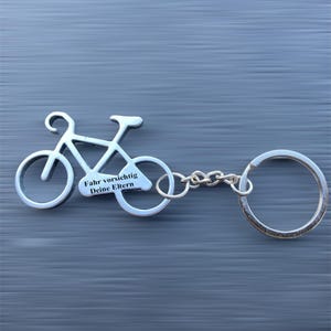 Könnte beinhalten: Silberfarbener Fahrrad-Schlüsselanhänger. Der Schlüsselanhänger zeigt eine Fahrradkontur mit den Worten "Fahr vorsichtig Deine Eltern" in weißer Schrift. Der Schlüsselanhänger ist an einer Kette und einem großen Schlüsselring befestigt.