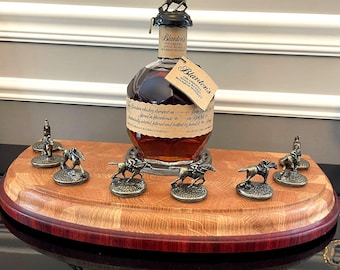 Puede incluir: Una botella de whisky bourbon Blanton's con un tapón de metal con un caballo y un jinete. La botella se encuentra sobre un expositor de madera con figuras de caballo y jinete de metal. El expositor tiene un borde rojo oscuro.