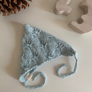 Puede incluir: Un gorro de bebé de punto azul claro con un lazo. El gorro tiene un patrón texturizado y una pequeña etiqueta blanca.