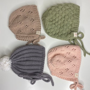May include: Four knitted baby bonnets in different colors and styles. The bonnets are made of soft yarn and have ties to secure them under the chin. One bonnet has a white pom-pom on top.