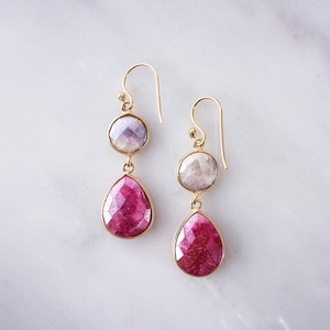 Könnte beinhalten: Goldene Ohrringe mit zwei tropfenförmigen rosafarbenen Edelsteinen und einem runden, hellvioletten Edelstein.