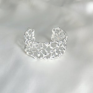 Op de afbeelding: Een zilveren armband met een delicaat, bladerig ontwerp. De armband is open en heeft een eenvoudige, elegante stijl.