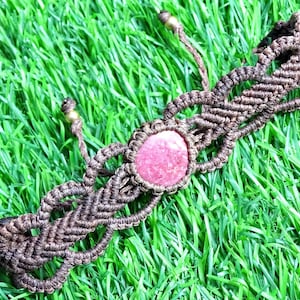 May include: A brown macrame bracelet with a pink stone in the center. The bracelet is made with a knotting technique and has a simple design.