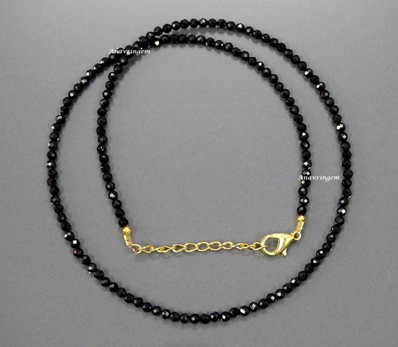 May include: A black beaded necklace with a gold chain extender. The necklace is made of small, round beads and has a simple, elegant design.
