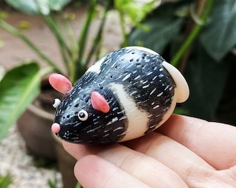 May include: A small, black and white ceramic mouse figurine with pink ears and a black nose. The mouse is sitting on a hand with the text "Miniclaysucculent" visible in the bottom right corner.