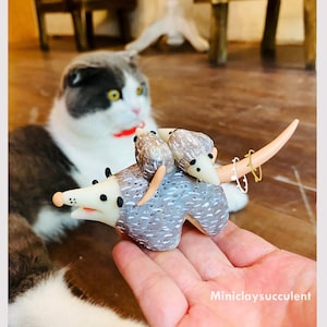 Könnte beinhalten: Eine handgefertigte Armadillo-Figur aus Polymer Clay mit einem gemusterten Panzer in Grau und Weiß. Das Armadillo hat einen langen Schwanz und trägt zwei kleinere Armadillos auf seinem Rücken. Die Figur sitzt auf der Hand einer Person.