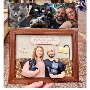 May include: A custom-made clay sculpture of a couple with their two cats. The sculpture is framed in a brown wooden frame and includes a plaque with the text "Our First Little Family Muty, Matt, Pees, Fancy & Selame Est. 2021".