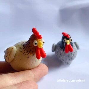 Peut inclure: Deux poulets en p&acirc;te polym&egrave;re faits &agrave; la main. L'un est brun et blanc avec une cr&ecirc;te et une barbe rouges. L'autre est gris avec une cr&ecirc;te et une barbe noires.