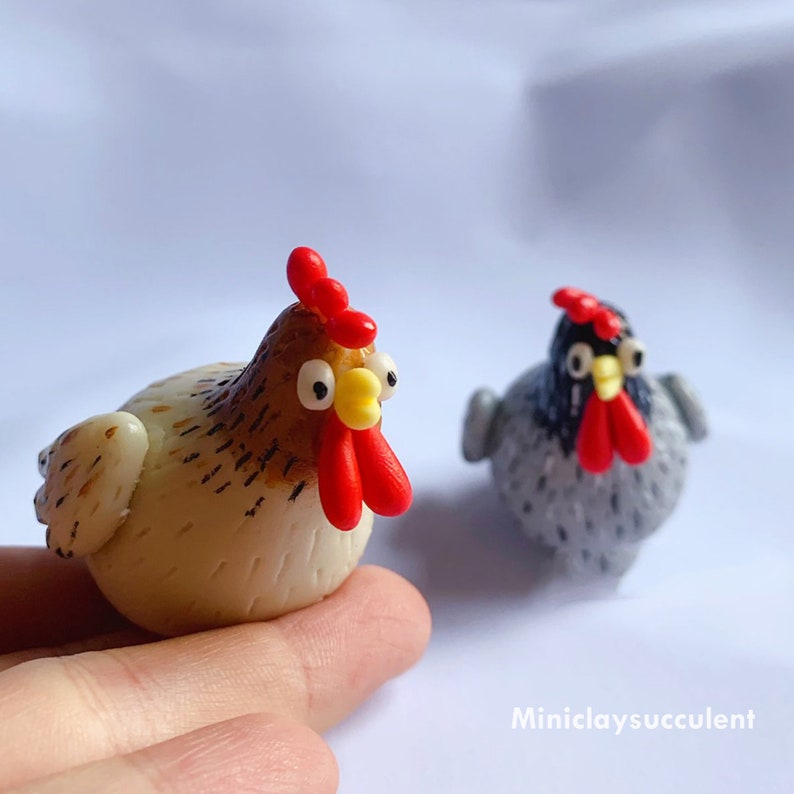 Peut inclure: Deux poulets en p&acirc;te polym&egrave;re faits &agrave; la main. L'un est brun et blanc avec une cr&ecirc;te et une barbe rouges. L'autre est gris avec une cr&ecirc;te et une barbe noires.