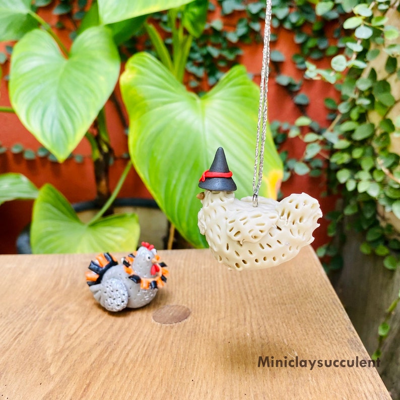 May include: A white ceramic chicken wearing a black witch hat with a red band, hanging from a silver string. A smaller gray ceramic chicken is sitting on a wooden surface.