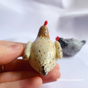 Peut inclure: Une figurine de poulet en c&eacute;ramique blanche avec des marques brunes et noires. Le poulet a une cr&ecirc;te rouge et un bec blanc. La figurine est pos&eacute;e sur une surface blanche.