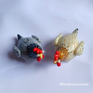 Peut inclure: Deux poulets en argile polym&egrave;re faits &agrave; la main, l'un gris avec des marques noires et l'autre blanc avec des marques brunes. Les deux poulets ont des cr&ecirc;tes et des barbillons rouges, des becs jaunes et des yeux noirs.