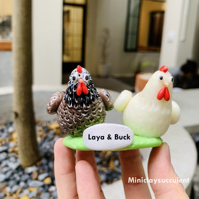 Peut inclure: Deux poulets en argile faits &agrave; la main, l'un brun et noir, l'autre blanc, avec des cr&ecirc;tes et des barbillons rouges. Ils sont assis sur une base verte avec une pancarte blanche qui indique "Laya & Buck".